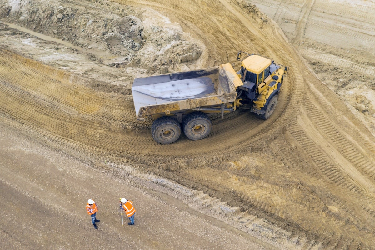 Mining Truck With Exposed Mining Truck Muffler On A Dirt Path, While Two Workers In Safety Vests Assess The Construction Site.